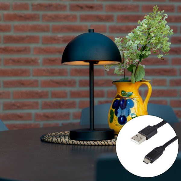 Schwarze Tischleuchte mit kuppelförmigem Schirm und USB-Ladekabel auf einem Tisch neben einer Vase vor einer Ziegelwand.