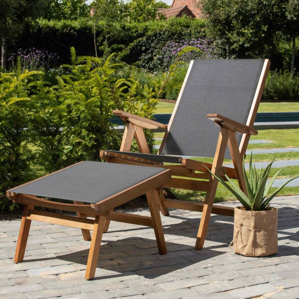 Moderner Garten-Loungesessel aus Holz mit Hocker und grauem Textilbezug auf einer gepflasterten Terrasse im sonnigen Garten.
