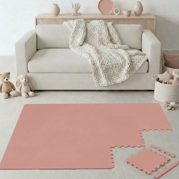 Rosa Schaumstoff-Puzzlematte im hellen Raum mit Sofa, Strickdecke und Stofftieren. Modulare Spielmatte aus ineinandergreifenden Teilen.