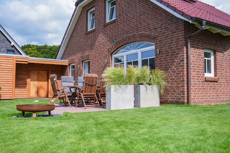 Gartenterrasse mit Holz-Gartengarnitur, grauen Sitzkissen, großen quadratischen Pflanzkübeln in Betonoptik und einer Feuerschale auf dem Rasen.