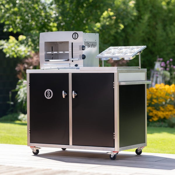 Beefer Oberhitzegrill auf einem schwarzen Outdoor-Küchenwagen aus Edelstahl mit Gastronorm-Behältern im Garten.