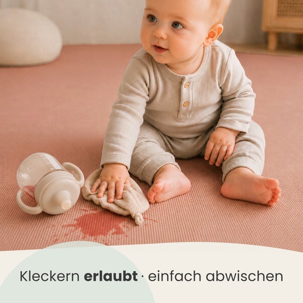 Ein Kleinkind sitzt auf einer abwischbaren Spielmatte und wischt mit einem Tuch einen verschütteten Fleck neben einer Schnabeltasse weg.