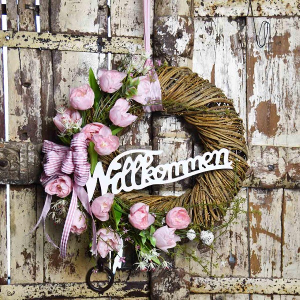 Dekorativer Türkranz mit rosa Blüten und der Aufschrift Willkommen an einer alten Holztür.