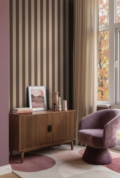Wohnzimmer mit gestreifter Tapete, Holzkommode, gepolstertem Sessel und gemustertem Teppich bei Tageslicht.