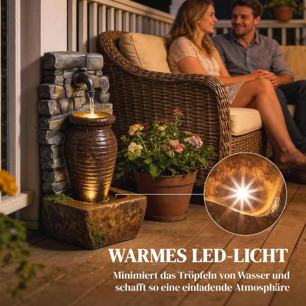 Gartenbrunnen in Steinoptik mit Amphore und LED Beleuchtung auf einer Terrasse neben einem Pflanzgefäß und Gartenmöbeln aus Geflecht.