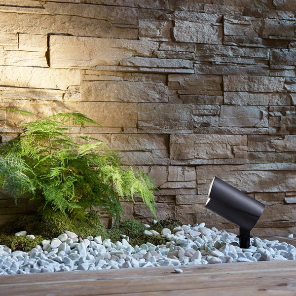 Schwarzer LED Gartenstrahler mit Erdspieß beleuchtet eine Steinwand und eine Pflanze in einem Beet aus weißen Kieselsteinen und Moos.