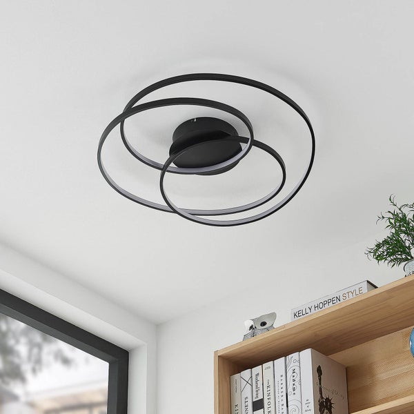 Moderne schwarze LED Deckenleuchte mit drei ineinander verschlungenen Ringen an einer hellen Decke über einem Holzregal.