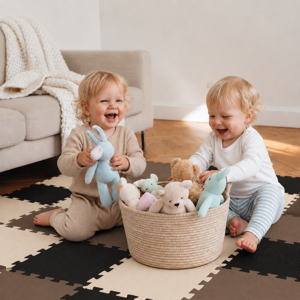 Zwei Kleinkinder sitzen auf einem Puzzle Spielteppich in einem Wohnzimmer und spielen mit Plüschtieren aus einem geflochtenen Korb.