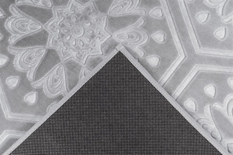 Grauer Teppich mit geprägtem Ornamentmuster und umgeklappter Ecke, die die rutschfeste Rückseite zeigt.