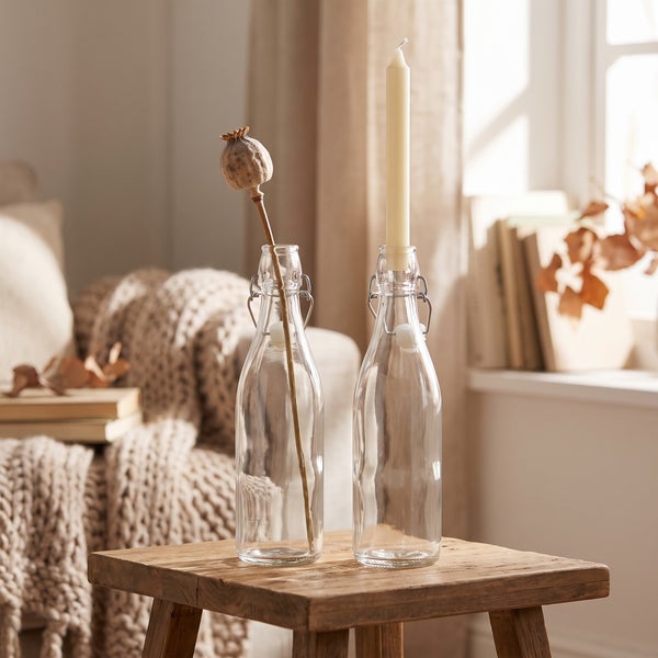 Zwei durchsichtige Glasflaschen mit Bügelverschluss auf einem kleinen Holztisch als dekorative Kerzenhalter und Blumenvasen.