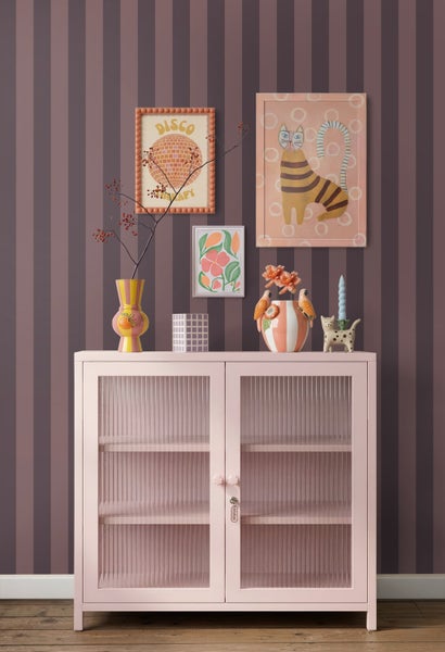 Rosa Glasschrank mit geriffeltem Glas vor gestreifter Tapete mit bunten Vasen und gerahmten Bildern auf einem Holzboden.