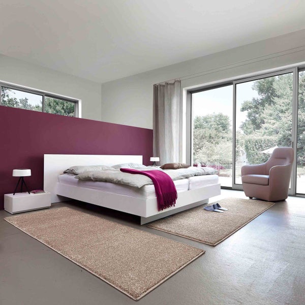 Helles Schlafzimmer mit weißem Bett, zwei beigefarbenen Teppichläufern auf grauem Boden, lila Akzentwand und Glasfront zum Garten.