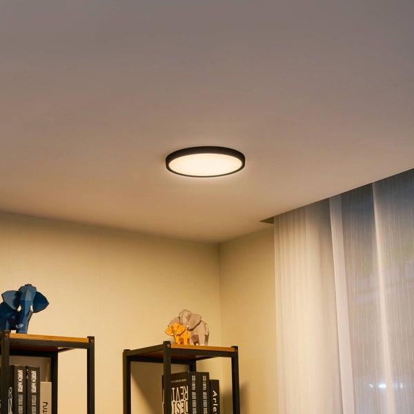Moderne runde LED-Deckenleuchte mit schwarzem Rahmen an einer Zimmerdecke in einem hellen Raum mit Regalen und Dekofiguren.