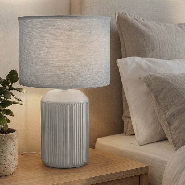 Moderne Tischleuchte mit geriffeltem Keramikfuß und Stoffschirm auf einem Holznachttisch neben einem Bett mit Kissen.
