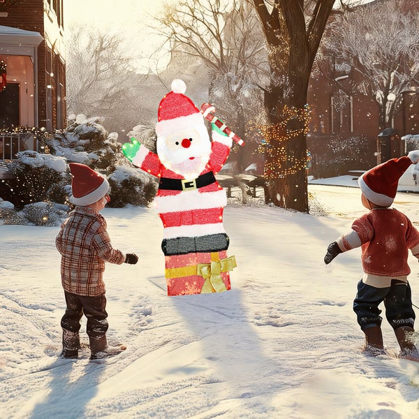Beleuchtete Weihnachtsfigur Weihnachtsmann stehend auf einem Geschenkpaket in einer winterlichen Gartenlandschaft mit zwei Kindern.