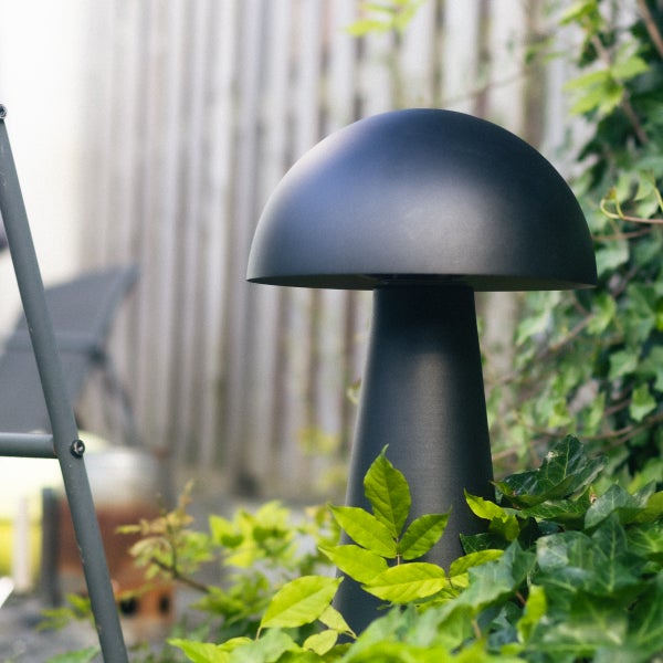 Moderne pilzförmige Außenstehleuchte aus schwarzem Metall in einem Garten zwischen Efeu und einem Holzzaun.