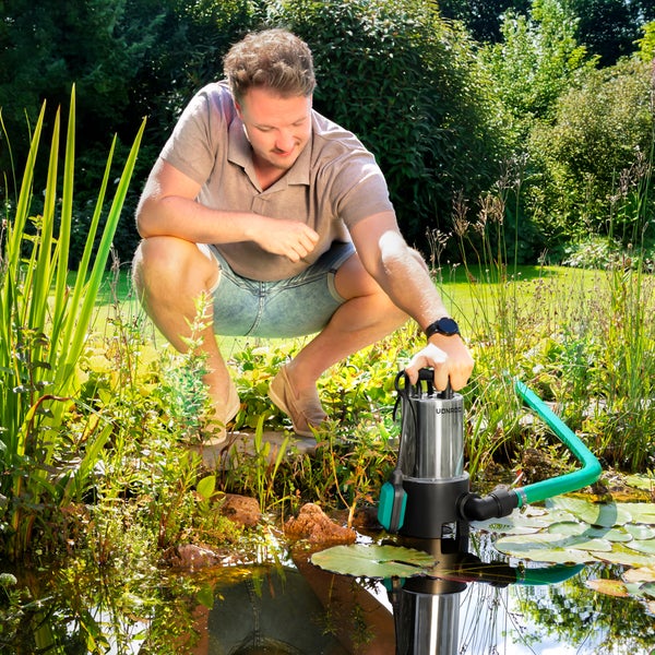 Ein Mann hockt an einem Gartenteich und setzt eine Edelstahl-Tauchpumpe mit einem grünen Schlauch in das Wasser neben Seerosenblätter.