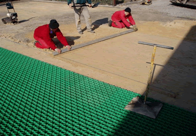 Arbeiter bereiten den Untergrund für Kunststoff-Rasengitter vor, indem sie das Sandbett mit einer Richtlatte ebnen.