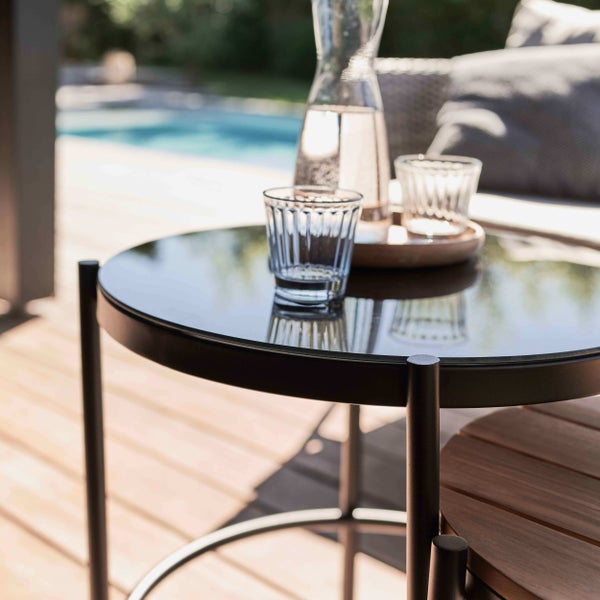 Runder Gartenbeistelltisch mit schwarzem Metallgestell und Glasplatte auf einer hellen Holzterrasse.