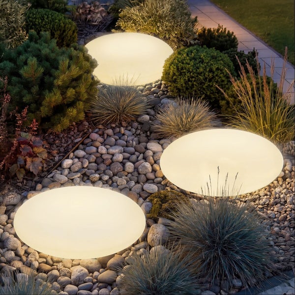 Drei hell leuchtende, ovale Außenleuchten in einem Gartenbeet mit Kieselsteinen und verschiedenen Zierpflanzen.