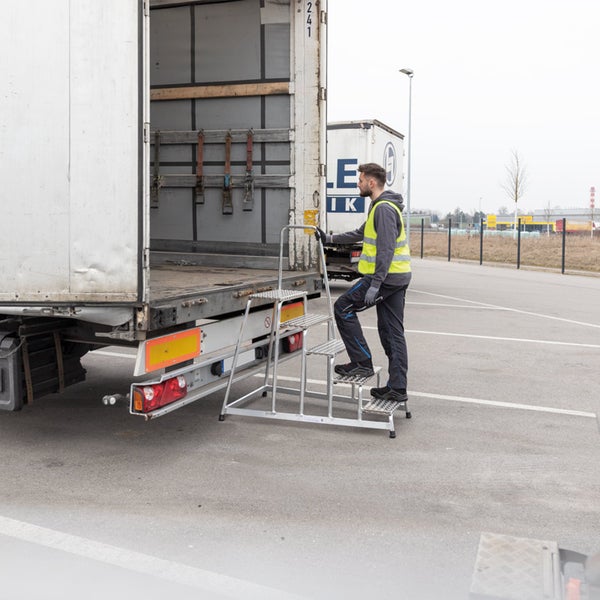 Arbeiter besteigt mit Hilfe einer Aluminium-Stufenleiter mit Handlauf einen LKW-Auflieger.