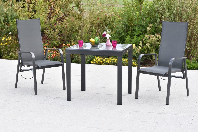 Gartenset mit zwei grauen Hochlehnern und einem quadratischen Glastisch auf einer hellen Steinterrasse vor einem blühenden Garten.