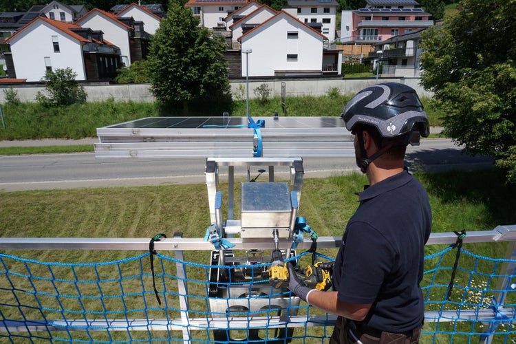 Arbeiter mit Schutzhelm bedient ein Hebegerät für Solarmodule mit einem Schlagschrauber in einem hellen Wohnviertel.