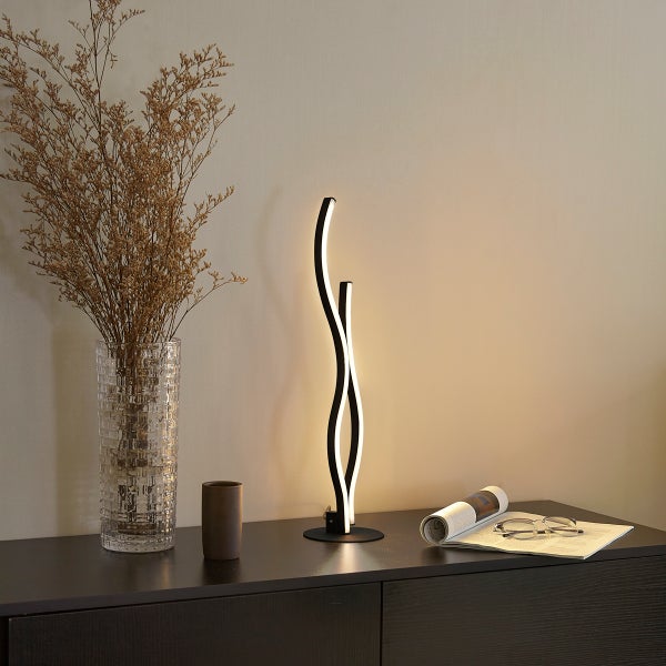 Moderne LED-Tischleuchte mit geschwungenem schwarzem Design auf einem dunklen Sideboard neben einer Glasvase mit Trockenblumen und einer Zeitschrift.