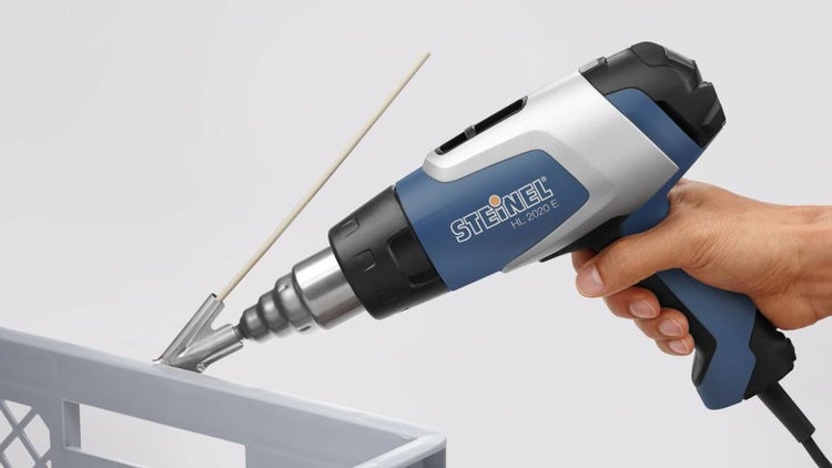 Hand führt Steinel Heißluftgebläse HL 2020 E mit Schweißdüse und Schweißdraht zum Kunststoffschweißen an einer Kunststoffkiste.