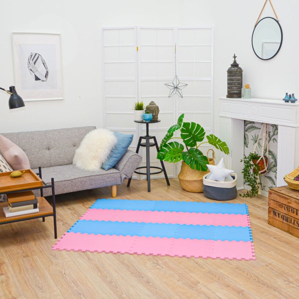 Helles Wohnzimmer mit blau-rosa Schaumstoff-Puzzlematte auf Holzboden. Modernes Design mit grauem Sofa, Monstera-Pflanze und Paravent.