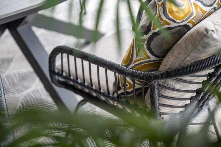Nahaufnahme eines modernen Gartenstuhls mit dunklen, geflochtenen Armlehnen, gemusterten und einfarbigen Kissen auf einer Terrasse.
