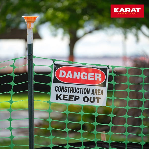Grüner Sicherheitszaun aus Kunststoff an einem Metallpfosten mit einem Warnschild für Baustellen. Karat Commercial Logo.