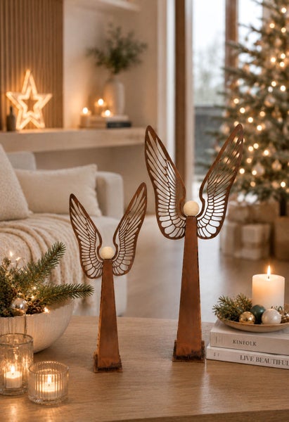 Zwei metallene Weihnachtsengel Figuren mit Flügeln und rundem Kopf auf einem Wohnzimmertisch vor weihnachtlicher Dekoration und Weihnachtsbaum.