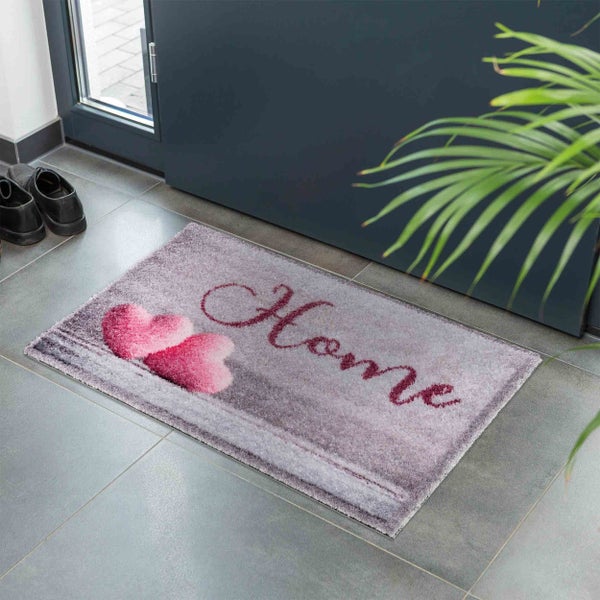 Fußmatte mit Herzmotiv und Schriftzug Home vor einer Haustür auf Fliesenboden