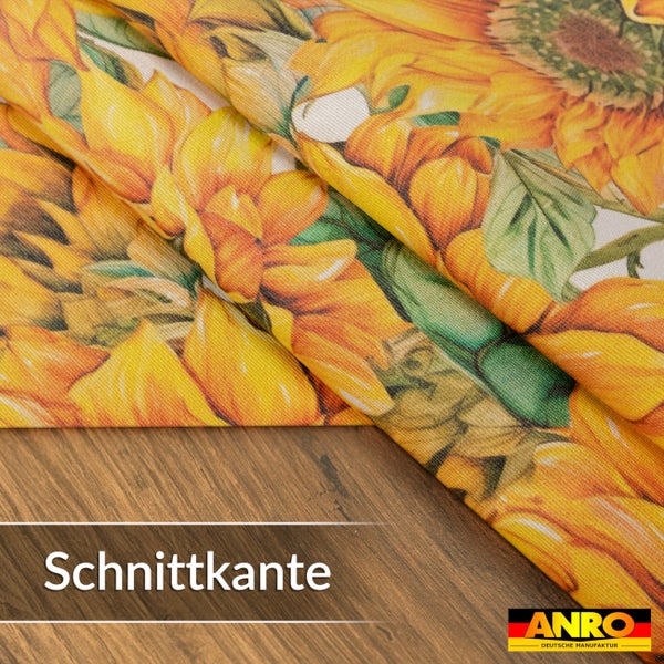 Nahaufnahme von Stoff Meterware mit Sonnenblumenmuster und einer Schnittkante auf einer Holzoberfläche. ANRO Logo.