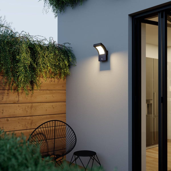 Moderne LED-Außenwandleuchte mit Bewegungsmelder an einer grauen Hauswand, Terrasse mit Holzlattenzaun, Pflanzen und schwarzen Gartenmöbeln.