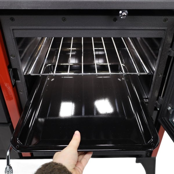 Hand zieht ein emailliertes Backblech aus dem Ofen eines Holzherdes mit darüber liegendem Grillrost.