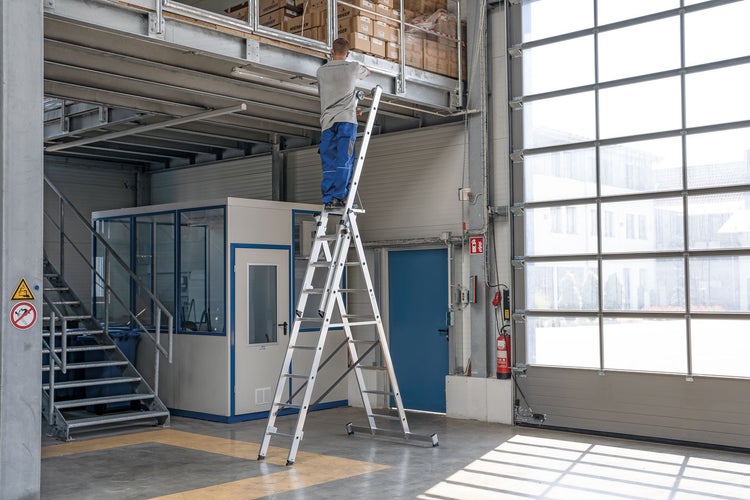 Mann auf einer Aluminium-Stufenleiter in einer Lagerhalle.