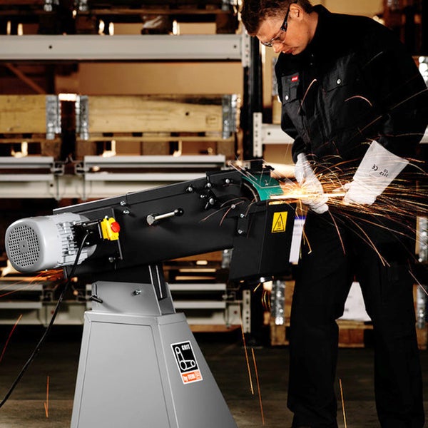 Ein Arbeiter in Schutzkleidung nutzt eine Bandschleifmaschine zur Metallbearbeitung in einer Werkstatt, während Funken fliegen.