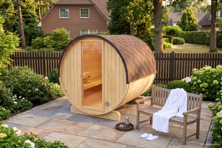 Eine Fasssauna aus Holz steht auf einer Steinterrasse in einem Garten, daneben befindet sich eine Holzbank mit einem Bademantel und Hausschuhen.