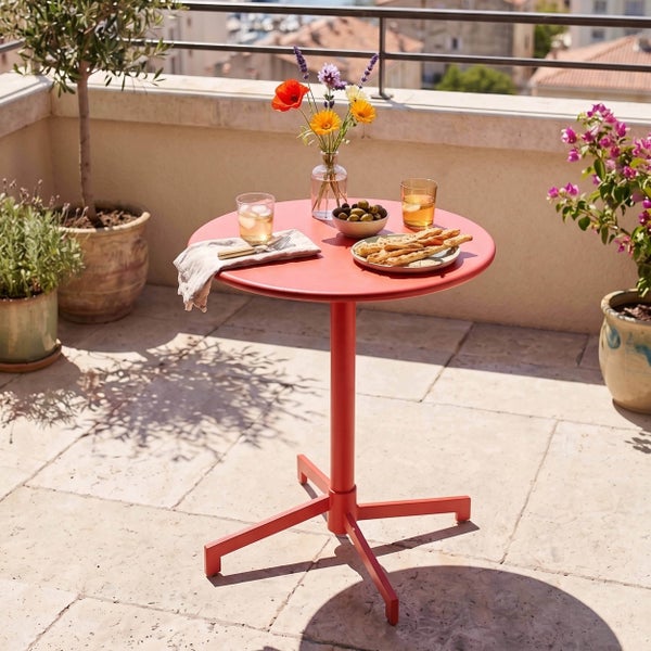 Runder Bistro-Tisch aus Metall auf sonnigem Balkon mit Steinfliesen, Blumen, Snacks und Getränken, umgeben von Topfpflanzen.