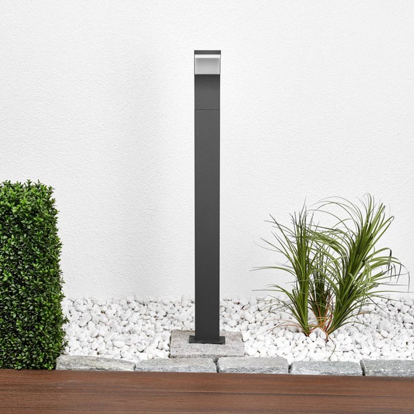Schlanke, schwarze Außenleuchte in einem hellen Garten mit weißen Steinen und Pflanzen vor einer weißen Wand.