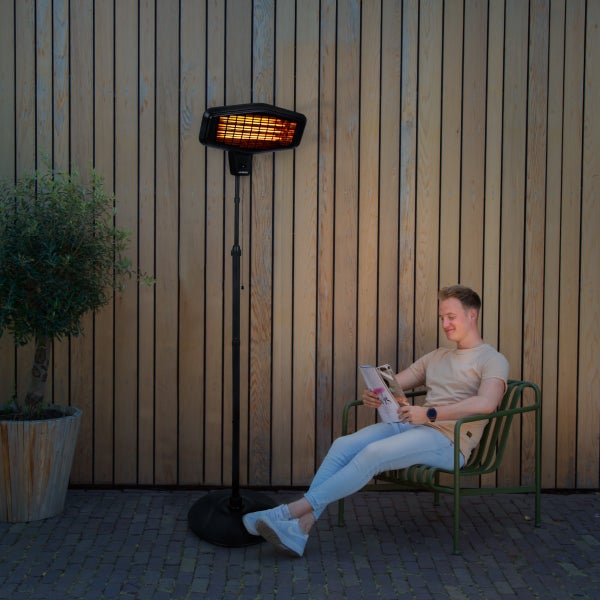 Elektrischer Terrassenstrahler auf Standfuß im Außenbereich neben einer sitzenden Person mit Magazin vor einer Holzwand und einer Pflanze.