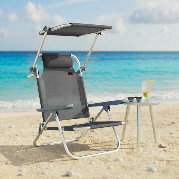 Klappbarer Strandstuhl aus Aluminium mit Sonnendach, Kopfstütze und Armlehnen an einem Sandstrand neben einem Beistelltisch mit Getränk.