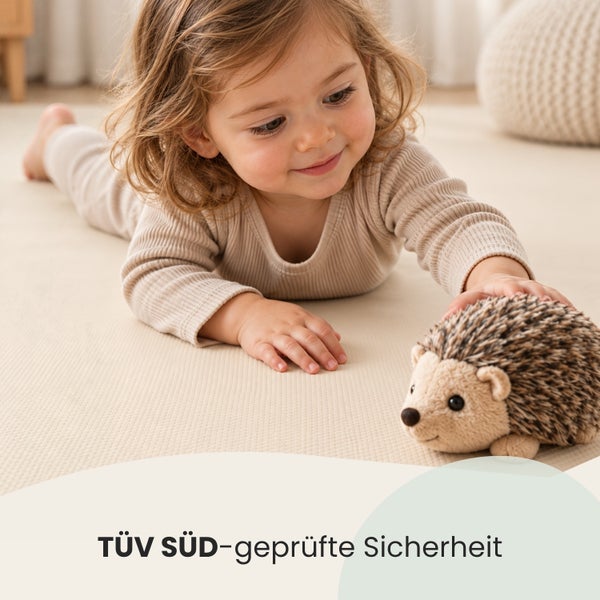 Ein Kleinkind spielt auf einem Boden mit einem Plüschigel, der das TÜV SÜD Siegel für geprüfte Sicherheit trägt.