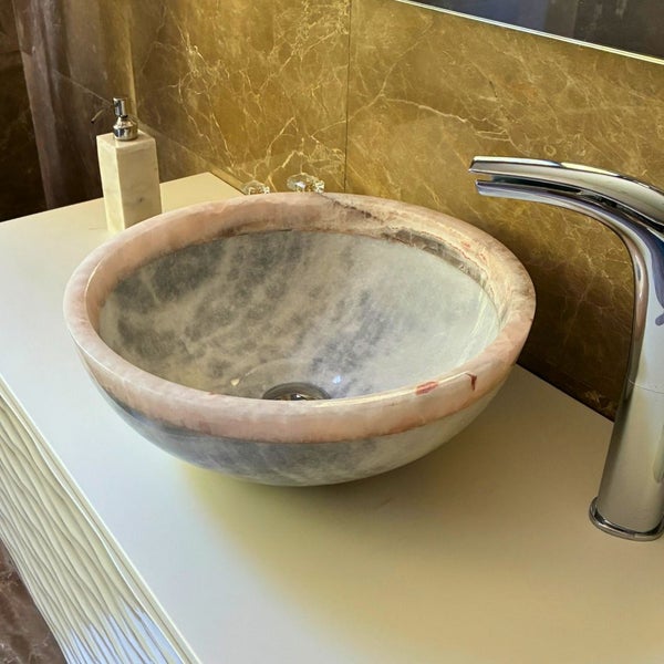 Rundes Aufsatzwaschbecken aus Naturstein auf einem hellen Waschtisch mit Armatur und Seifenspender vor einer marmorierten Wand.