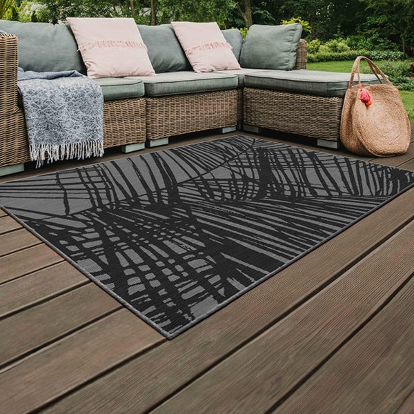 Rechteckiger Outdoor-Teppich mit Palmenblatt-Muster auf einer Holzterrasse mit einem Rattan-Ecksofa und Kissen.