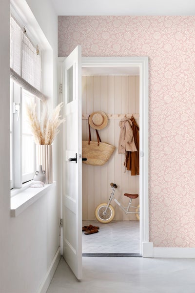 Hell eingerichtetes Zimmer mit rosa gemusterter Tapete, offener weißer Tür zum Flur mit Holzpaneelen, einer Strohtasche und einem Laufrad.