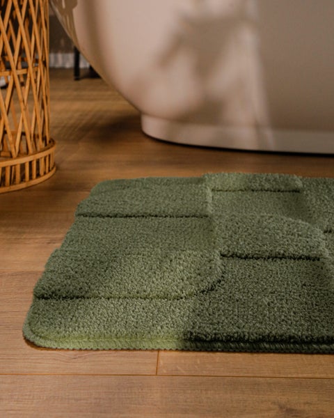Grüner Badevorleger mit geometrischem Karomuster auf Holzboden vor einer weißen Badewanne in einem hellen Badezimmer.