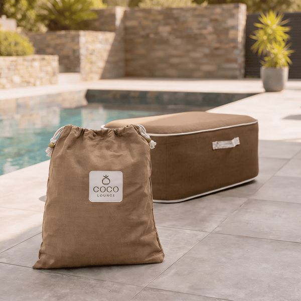 Brauner Coco Lounge Outdoor-Pouf mit weißer Paspelierung und passender Aufbewahrungstasche auf einer Steinterrasse am Pool.